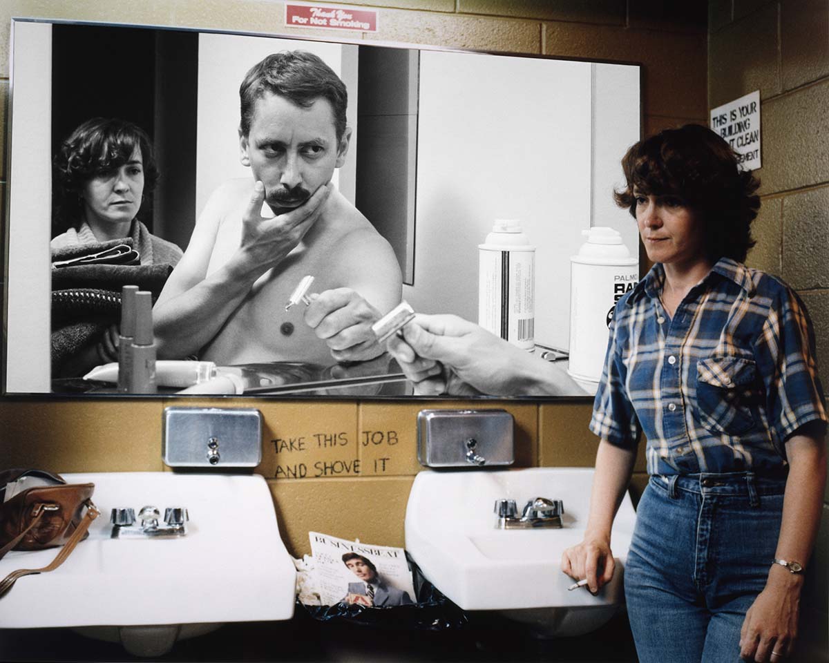 Photo d’une femme dans une toilette publique où le miroir a été remplacé par une photo en noir et blanc d’un homme se rasant. 