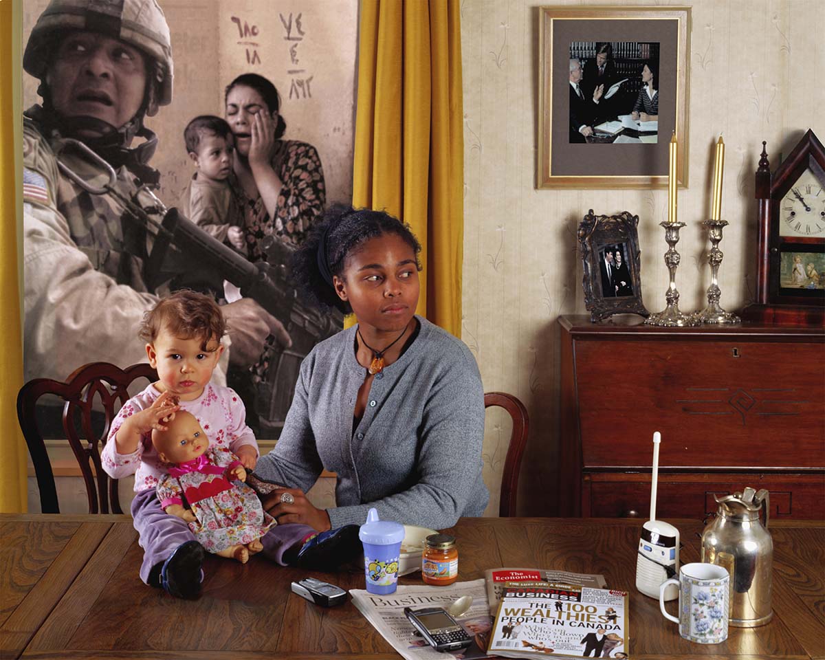Une femme assise à une table tenant un enfant avec une photo sur le thème de la guerre en arrière-plan.  