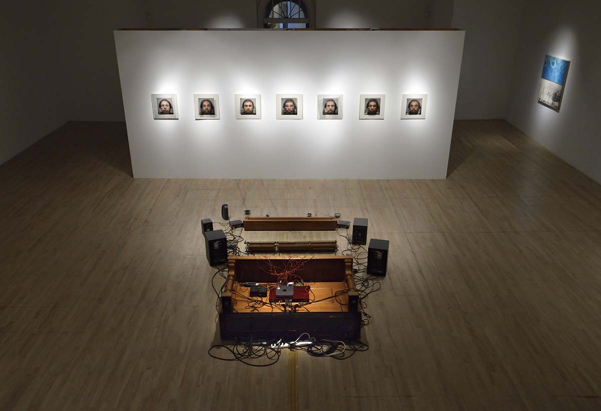 Une salle d’exposition montrant sept photos d’hommes sur un mur blanc placés en face d’un système de son. 