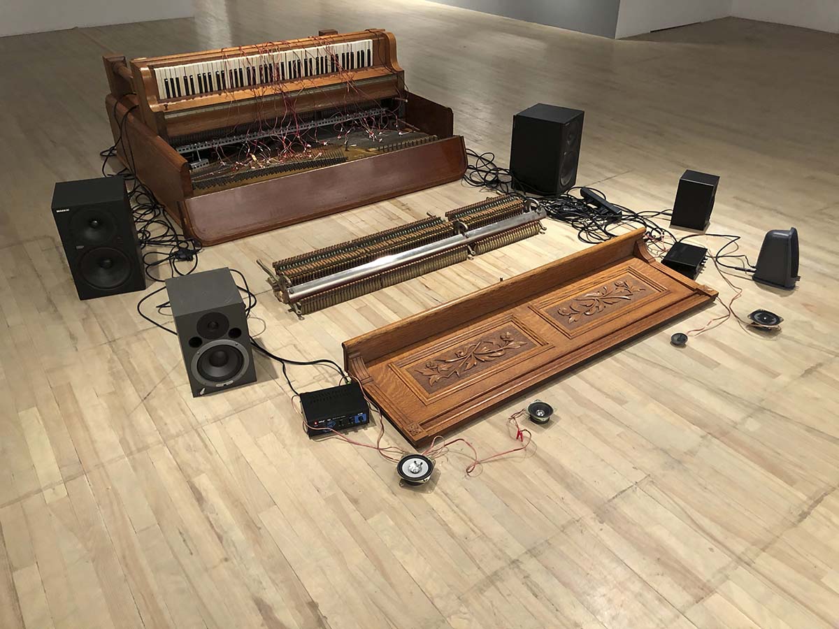 Installation montrant un piano désassemblé et un système de son posés sur le plancher d’une salle d’exposition. 