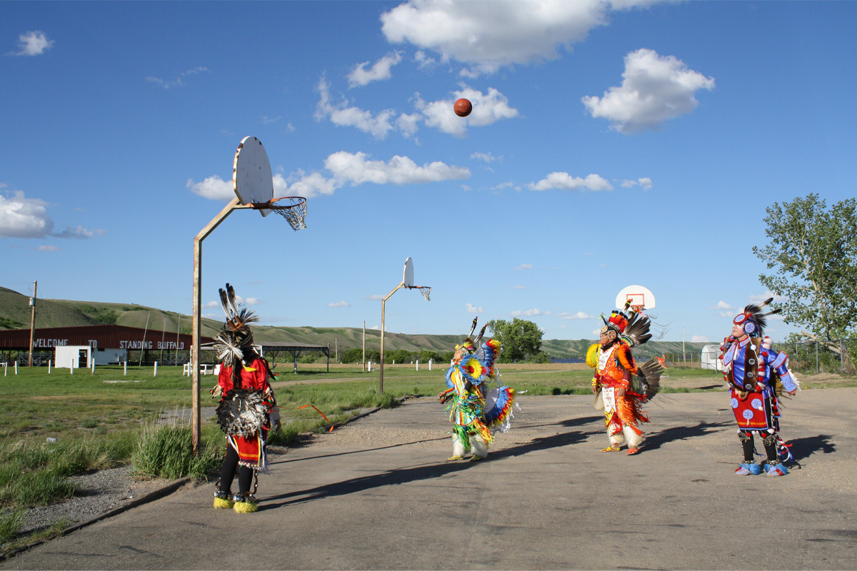 Quatre personnes sont sur un terrain de basketball, un ballon au-dessus de leurs têtes. Elles portent des coiffes à plumes et des tenues autochtones traditionnelles colorées et ornementées. 