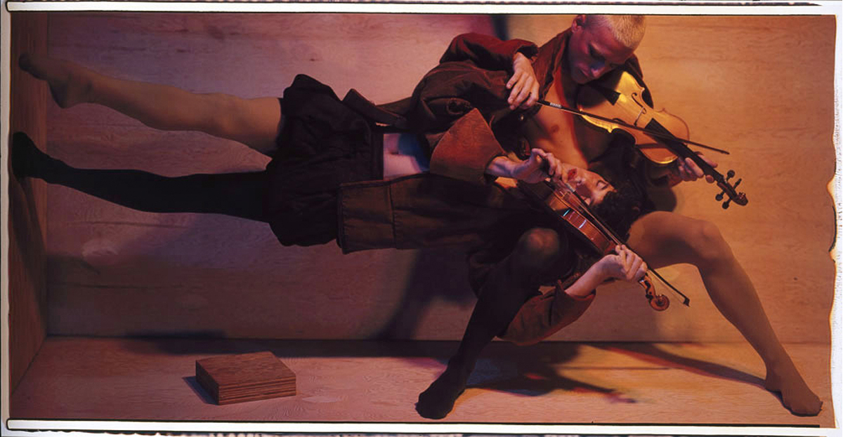 Un violoniste joue de son instrument, adossé contre un mur. Un deuxième violoniste, la tête posée sur les genoux de la première personne, se tient debout sur le plan horizontal, ses pieds posés contre un mur. 