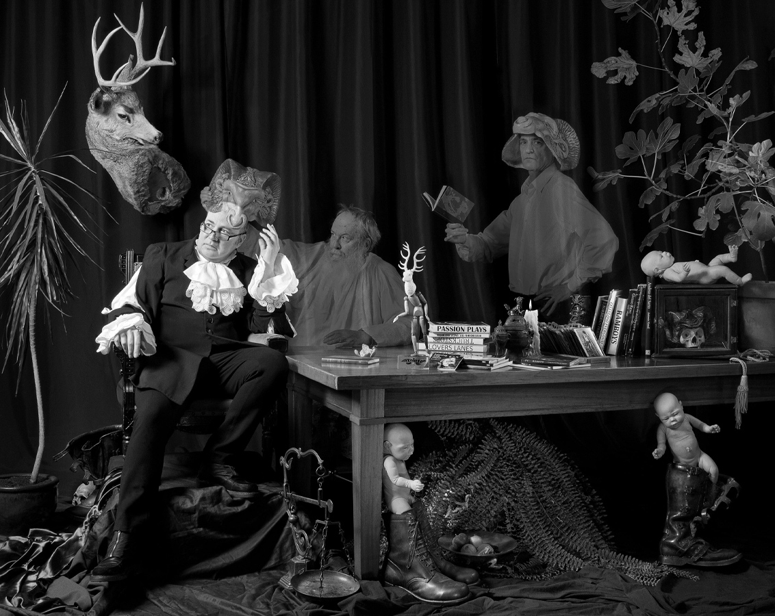 Un homme portant une tenue d’époque est assis, accoudé à une table. Divers objets l’entourent, dont des figurines de bébés et des silhouettes fantomatiques portant des masques de béliers. 