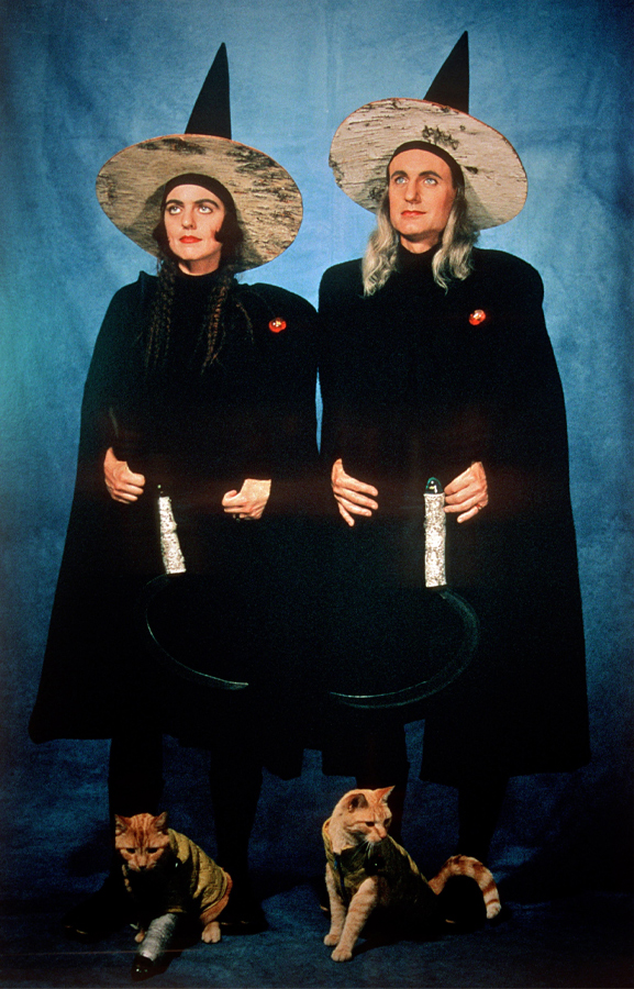 Portrait de deux personnes portant des robes noires et des chapeaux de sorcière, tenant des faux, et se tenant devant un arrière-plan bleu. À leurs pieds se tiennent deux chats roux. 