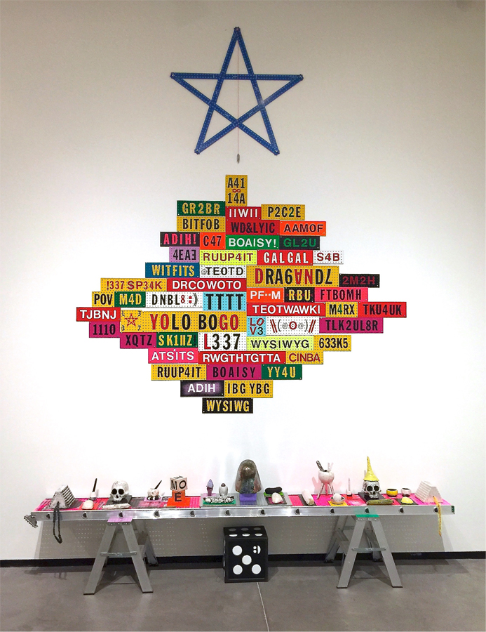 Objets symboliques disposés sur une échelle installée à l’horizontale. Au-dessus, le mur présente un montage d’affichettes rectangulaires multicolores garnies de sigles en lettres majuscules. 