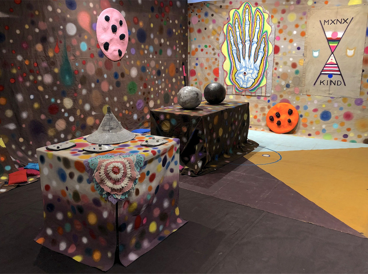 Des bâches couvertes de cercles colorés sont installées devant deux murs et sur deux tables. Divers objets sont dispersés, dont des sphères grises et un tableau illustrant le squelette d’une main. 