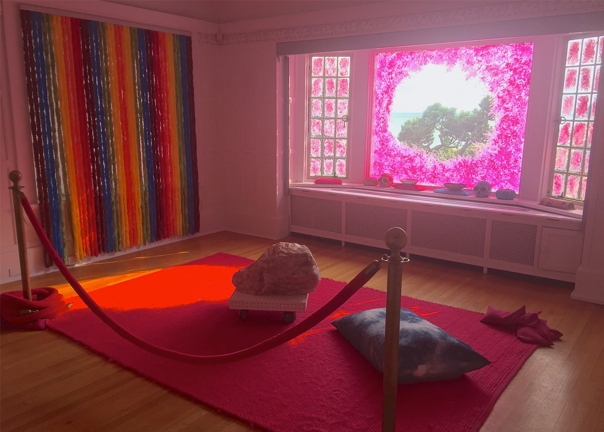 Une grosse pierre et un coussin sont posés sur un tapis rouge. Au fond, une fenêtre est tachetée de peinture rose, et au mur, un rideau multicolore de tubes en tissu formant un arc-en-ciel est suspendu. 