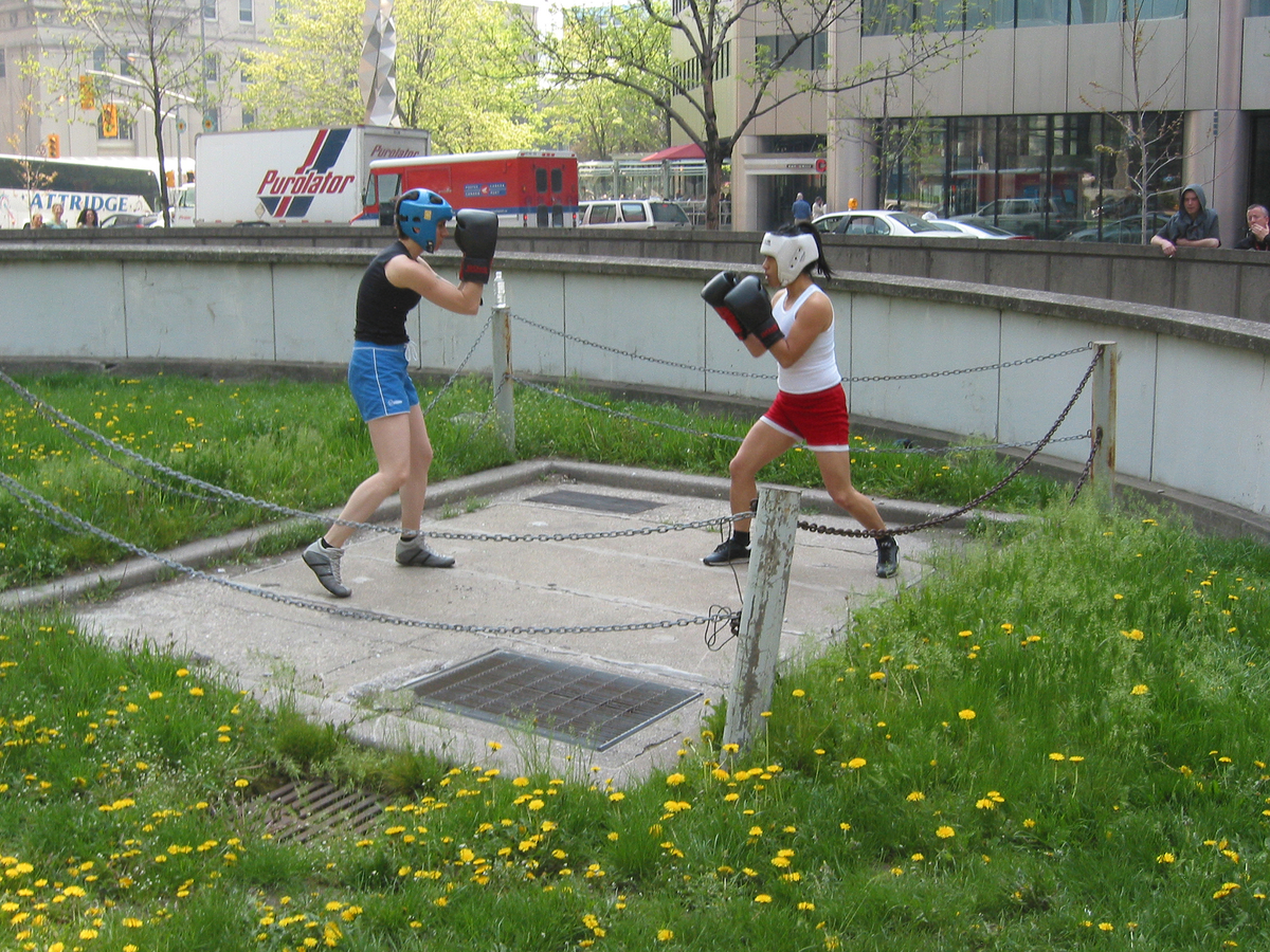 Deux jeunes portant des gants et des casques de boxe se livrent à un combat sur un espace bétonné, près d’une rampe d’accès à un stationnement sous-terrain.  