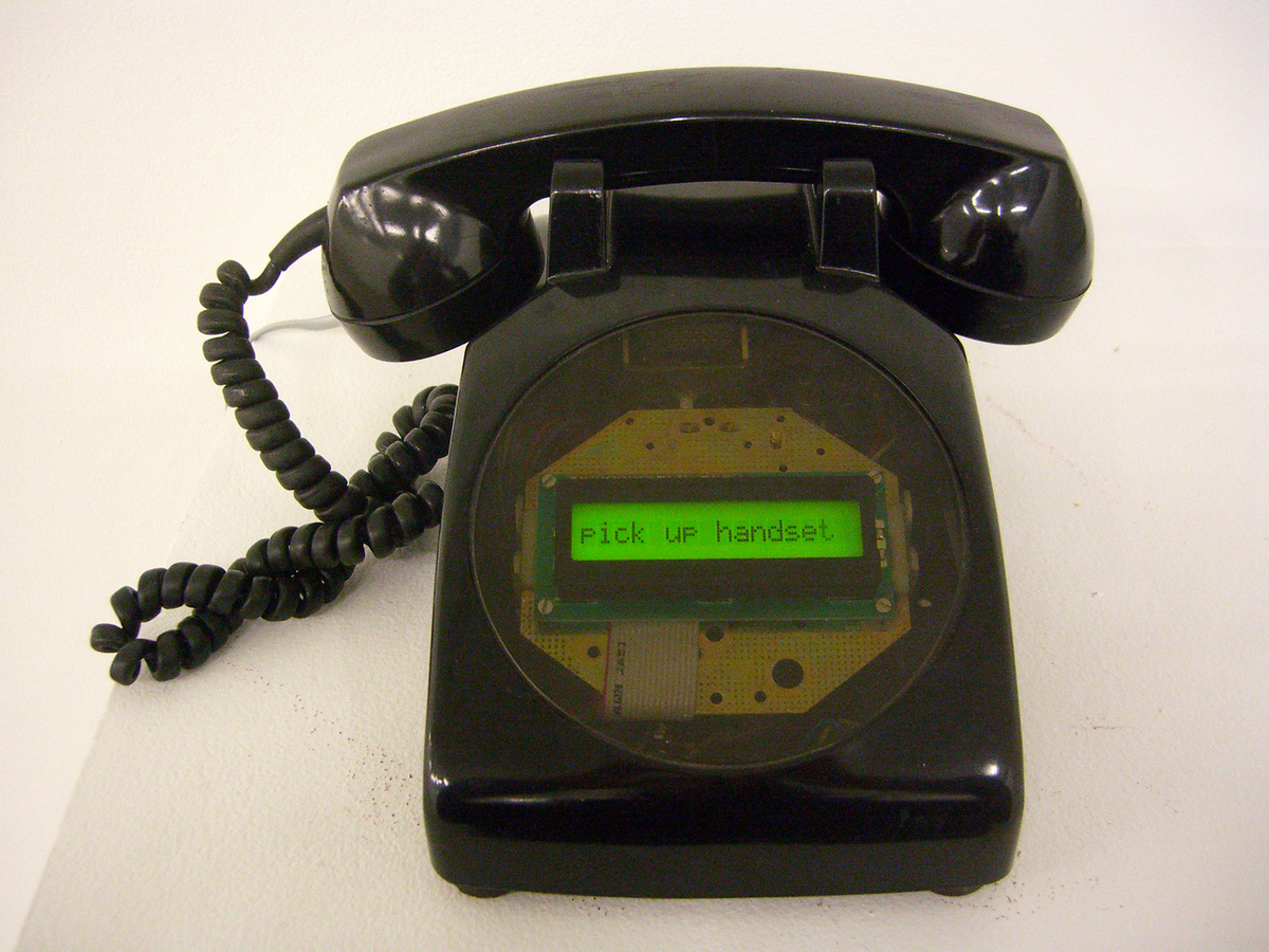 Un téléphone noir, qui date des années 1970 et qui est déposé sur une surface blanche, est muni d’un écran vert à DEL indiquant les mots « Pick up handset ». 