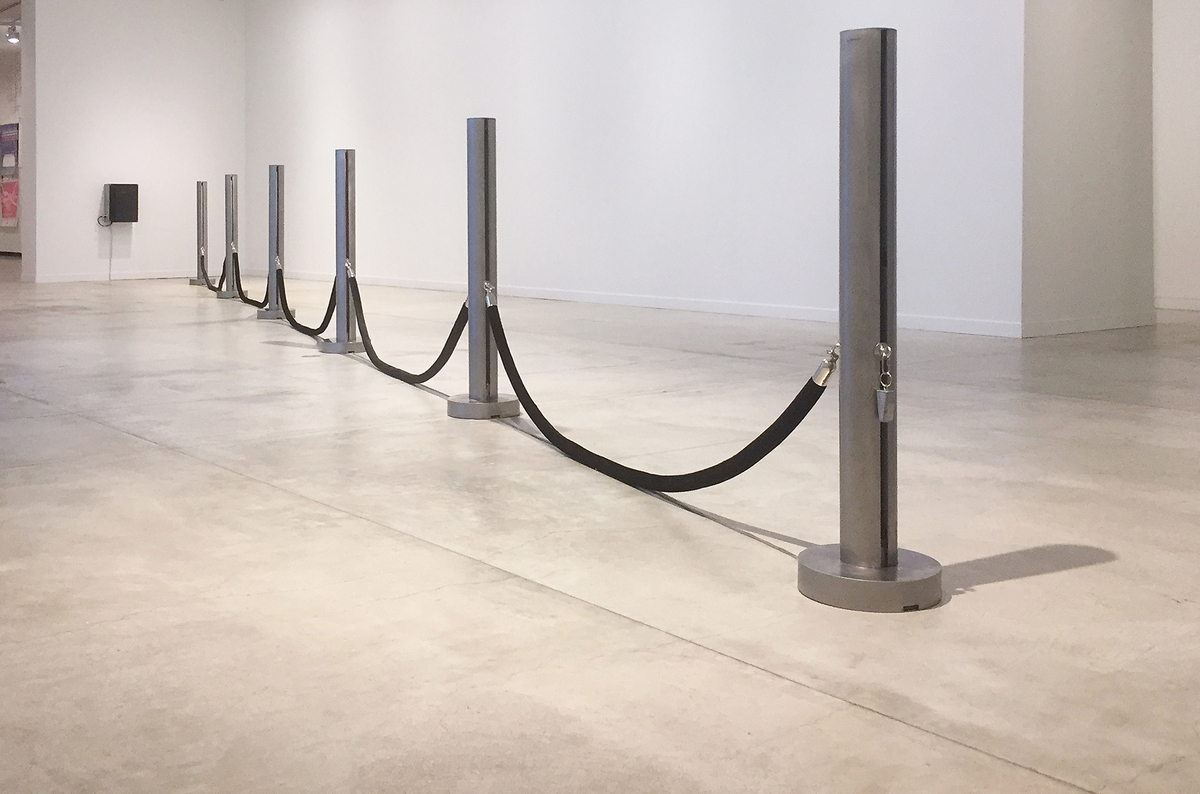 Des tubes de tissu noir sont suspendus entre des poteaux équidistants alignés dans une galerie, devant un casier noir monté au mur. 