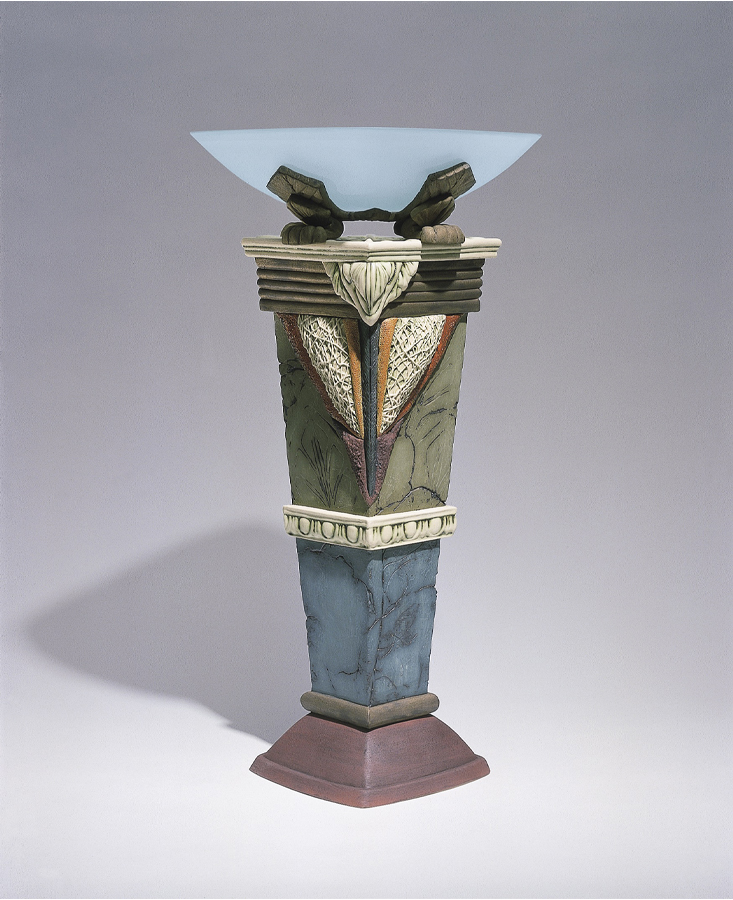 Grand bol évasé en verre translucide déposé sur un socle de céramique à quatre faces. Le vase est décoré de formes glacées dans des tons verts, bleus et rouges.  