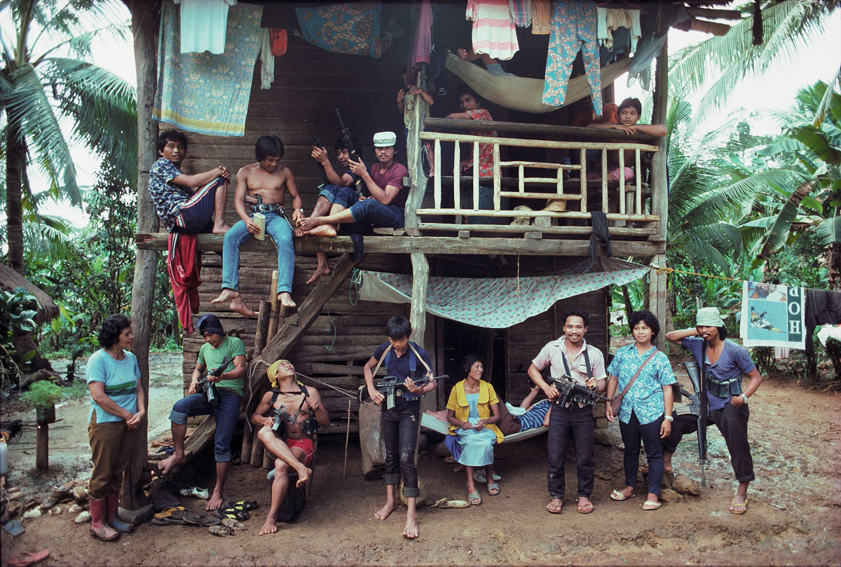 Des hommes, des femmes et des enfants − dont certains tiennent des fusils − posent devant et dans une structure de rondins à deux étages construite dans la jungle. 