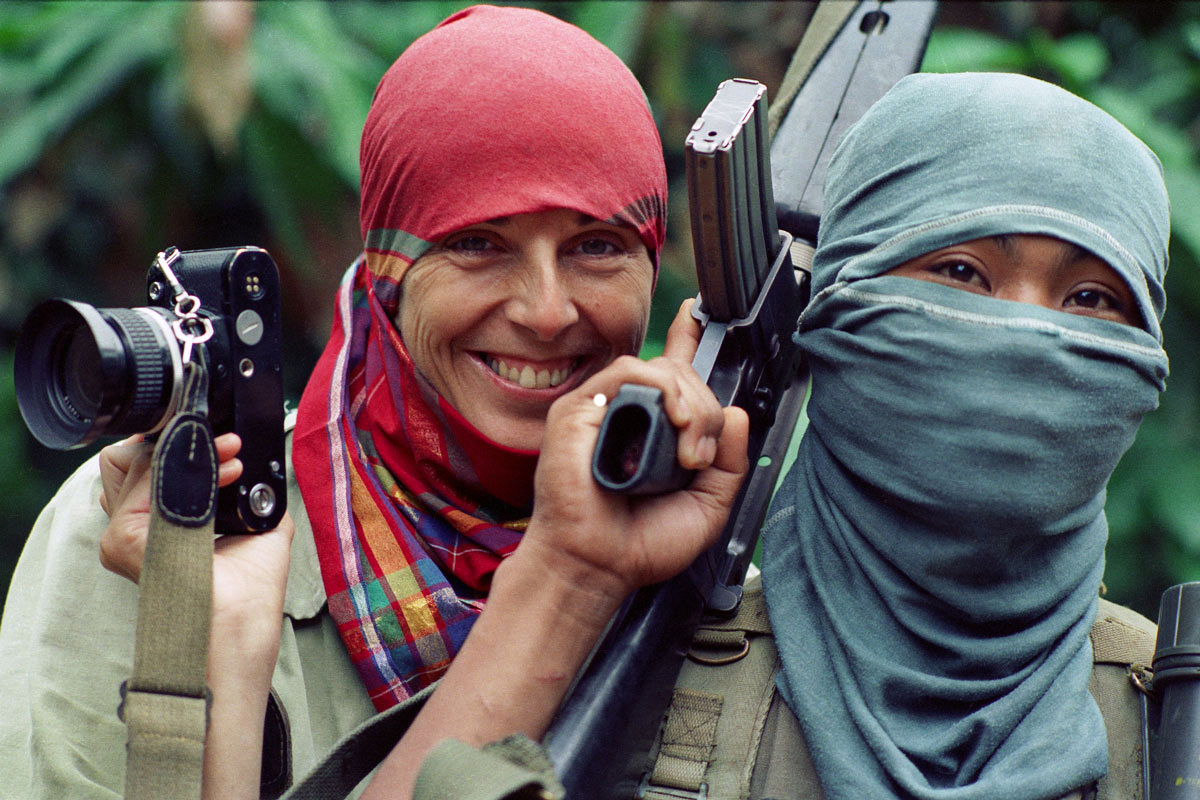 L’artiste souriante, coiffée d’un foulard rouge, tient un appareil photo près de sa tête. Elle se tient derrière un homme portant une cagoule et tenant un fusil d’assaut. 