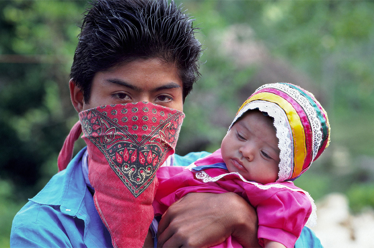 Un homme aux cheveux noirs et brillants et au le visage couvert d’un bandana tient un bébé portant une robe et un bonnet colorés. 