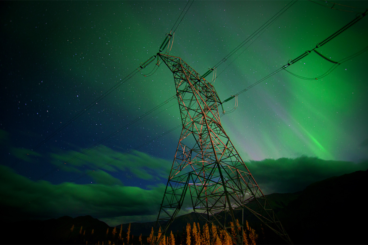 Un pylône hydroélectrique se découpe sur un ciel étoilé où miroitent des aurores boréales dans des tons flamboyants de vert, devant des montagnes ombragées. 