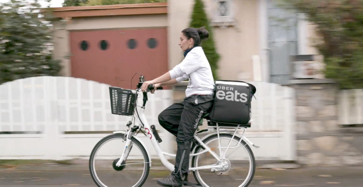 Une femme roule dans un quartier résidentiel sur un vélo équipé d’une boîte Uber Eats. Elle porte des boucles d’oreille, des pantalons imperméables et un chandail molletonné.  