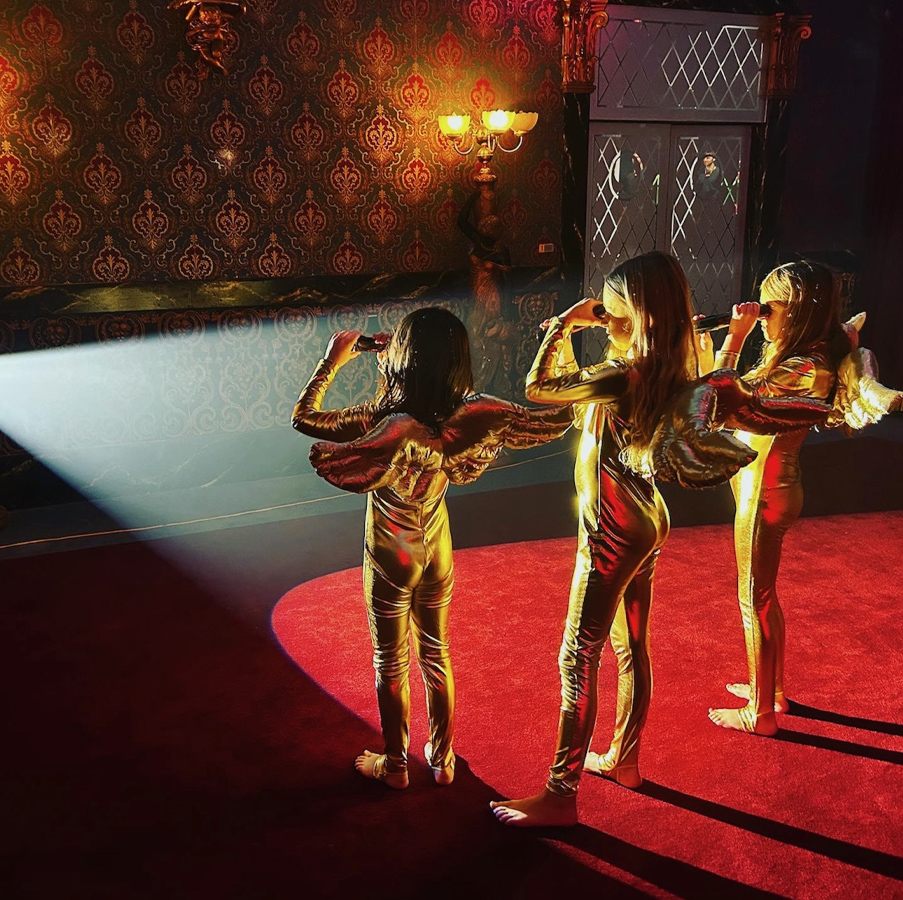 Trois fillettes sont debout sur le tapis rouge d’une scène, pieds nus, sous les projecteurs. Chacune porte une combinaison ailée de couleur or et regarde dans une lunette. 