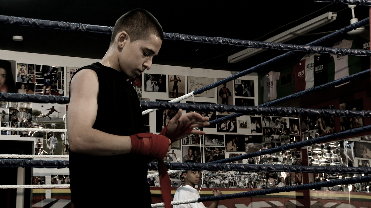 Un jeune homme est debout dans un ring de boxe, installant des bandages sur ses mains, devant un mur où sont affichés des tee-shirts et des photos d’athlètes.  