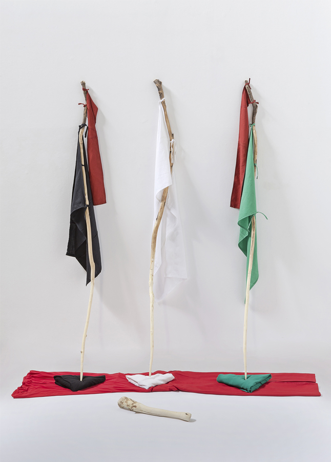 Morceaux de tissu blancs, noirs, rouges et verts attachés à trois bâtons appuyés contre un mur blanc. Les bâtons sont posés sur des morceaux de tissus des mêmes couleurs.  