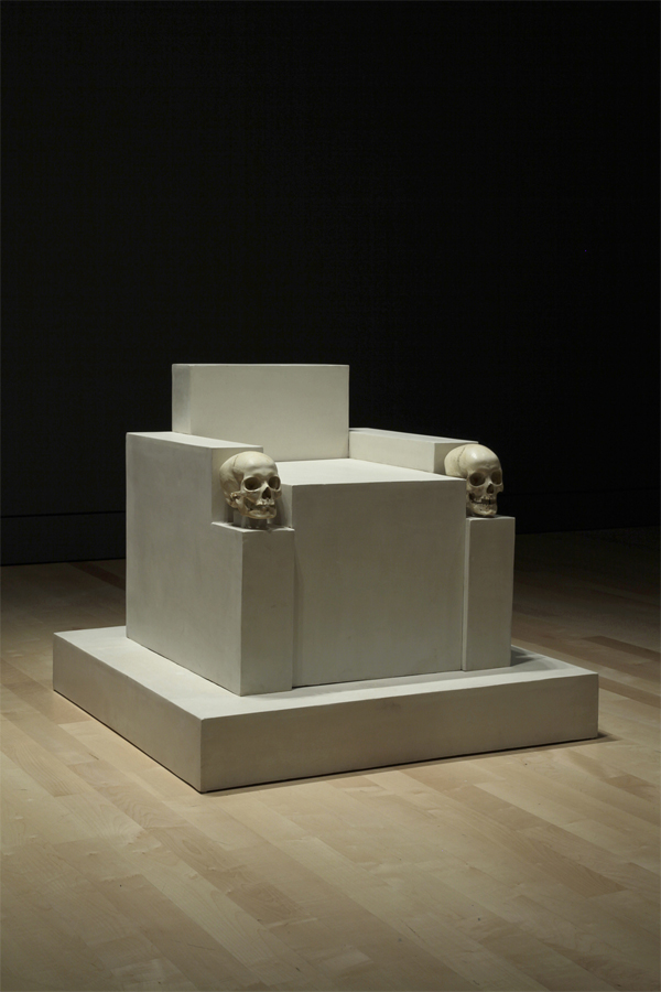 Un assemblage de volumes blancs suggérant un trône sobre au siège bas est déposé sur un socle assorti. Le devant des accoudoirs est décoré de crânes humains.  