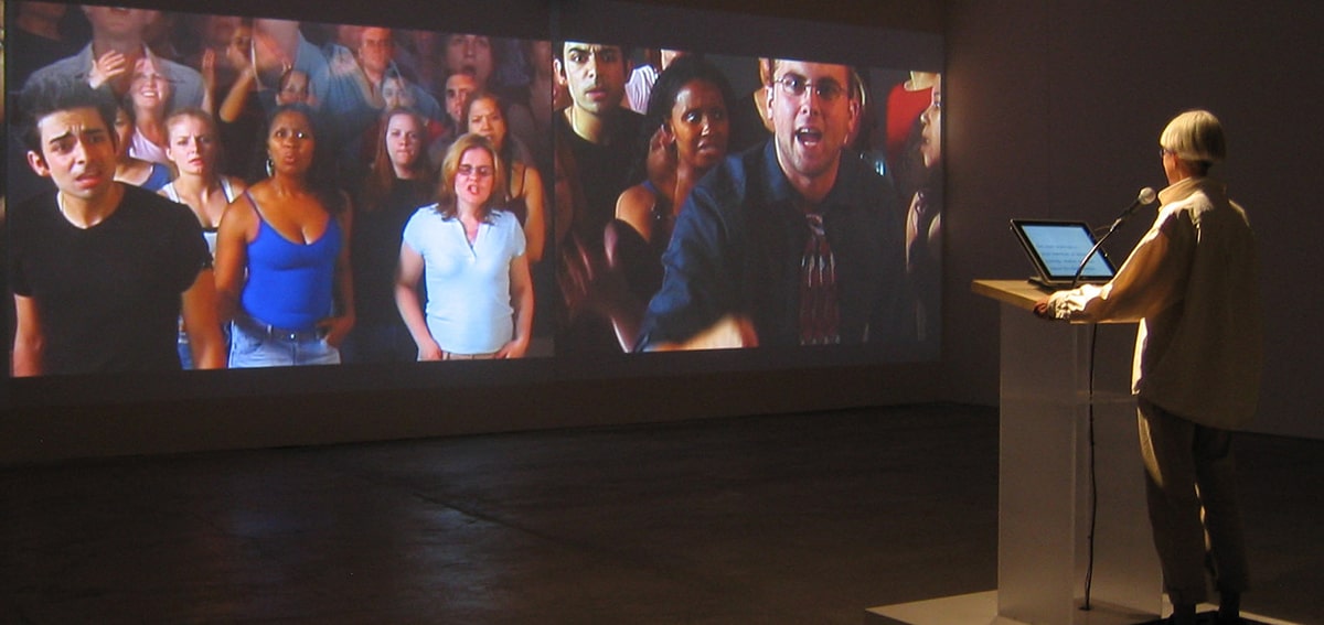 Une personne derrière un lutrin fait face à un écran sur lequel est présenté plusieurs autres personnes. 