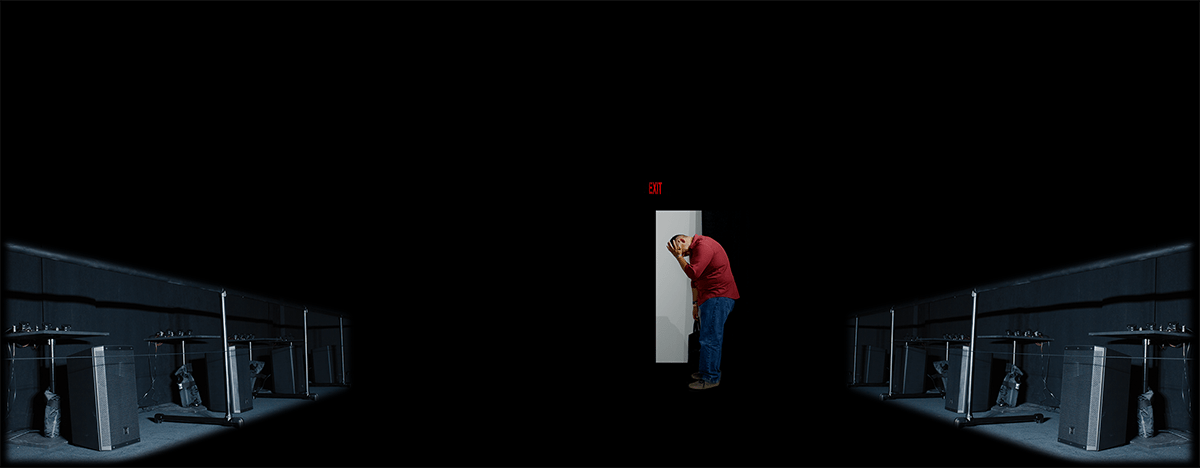 Un homme entouré de haut-parleurs se trouve devant une porte blanche dans une salle noire. 